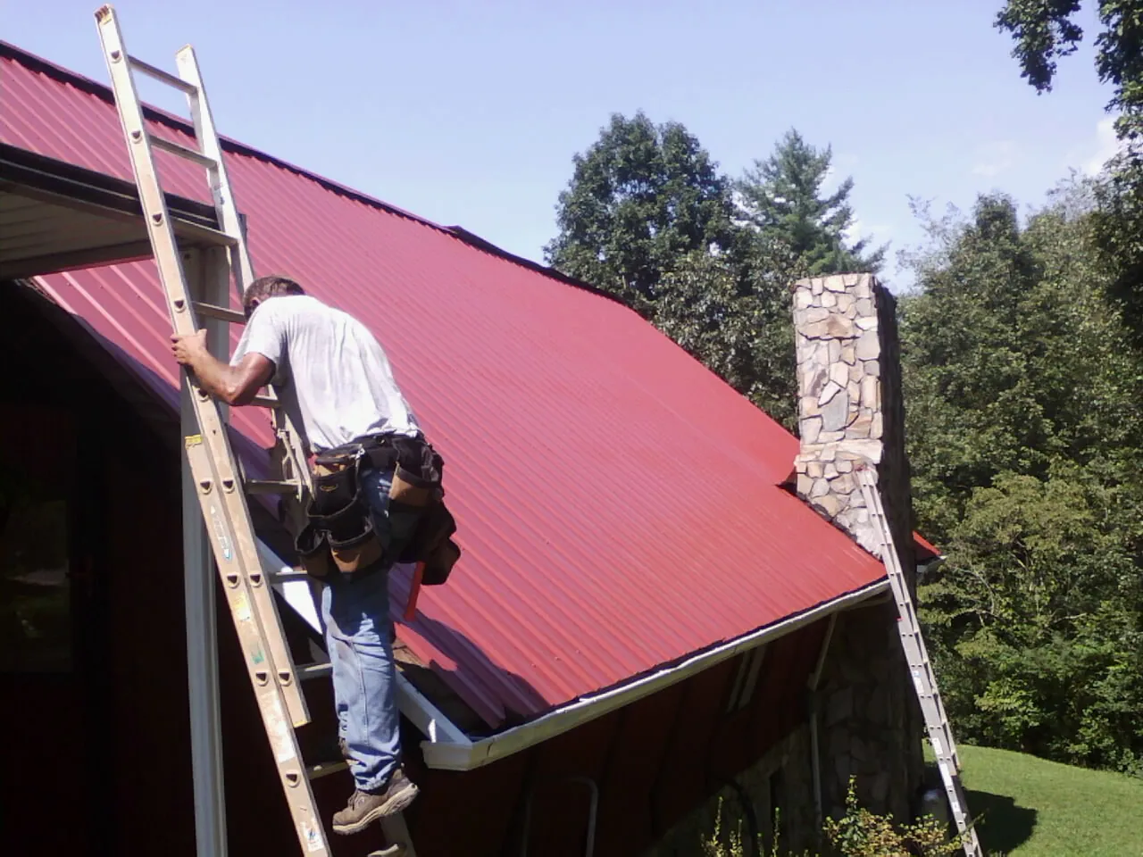 Roofer on a ladder beside a red metal roof and stone chimney — Soffit & Fascia Repair by Roofing Brothers in Lawrence Township